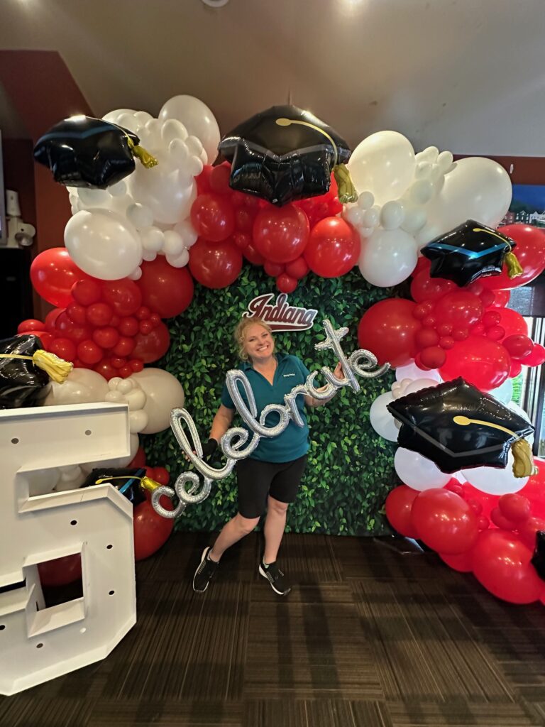 Custom graduation balloon backdrop with red and white balloon garland, black graduation cap balloons, silver “celebrate” script balloon, and greenery wall backdrop rental. Indiana University graduation celebration with luxury graduation balloons, graduation photo backdrop, and the best balloon decor in St. Louis. Graduation decor for parties, custom balloon installation, and greenery backdrop in St. Louis event venue.