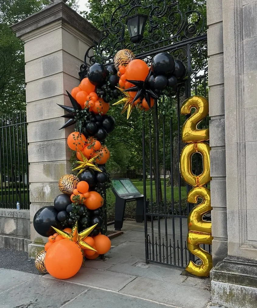 Graduation balloon garland on wrought iron gate with orange and black balloons, gold foil “2025” numbers, chrome starburst balloons, and tiger print accents. Custom balloon decor for Princeton University graduation. Outdoor graduation balloon installation, luxury graduation balloons, balloon arch for commencement, balloon garland for university events, best graduation balloons in Princeton and St. Louis.