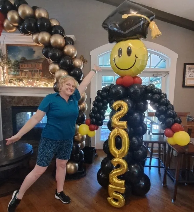 Life-sized graduation balloon sculpture of “Grad Guy” with smiley face balloon head, black balloon body, gold “2025” number balloons, and graduation cap foil balloon. Custom balloon decor for indoor graduation party with gold and black balloon arch. Fun graduation balloon ideas, balloon artist in St. Louis, graduation party decorations, best graduation balloons in the Greater St. Louis area.