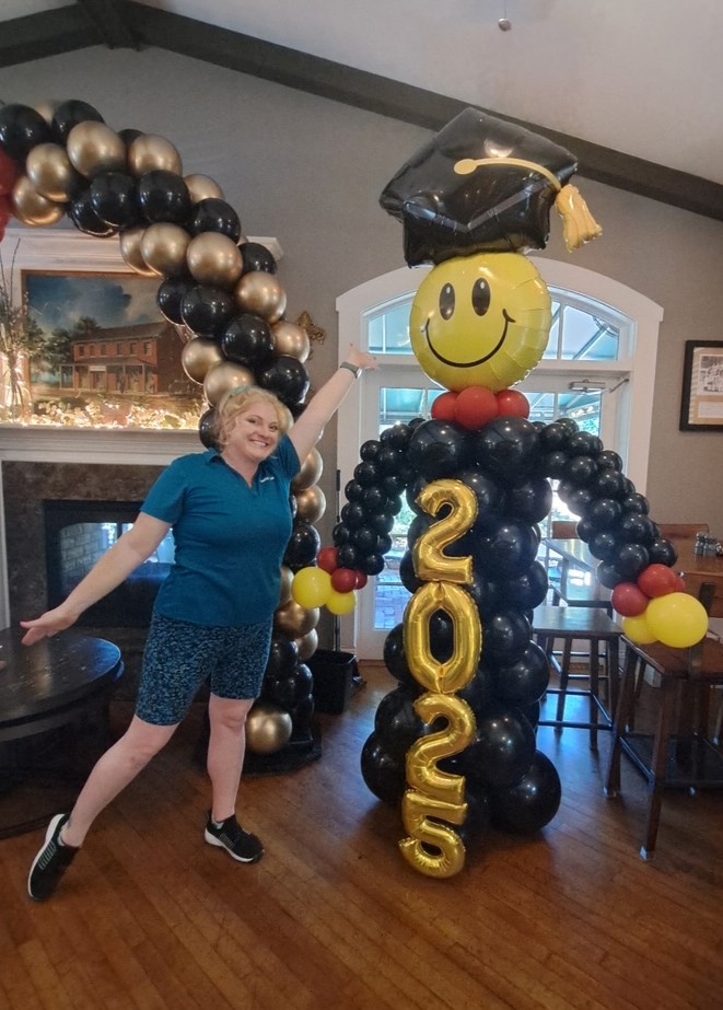 Life-sized graduation balloon sculpture of “Grad Guy” with smiley face balloon head, black balloon body, gold “2025” number balloons, and graduation cap foil balloon. Custom balloon decor for indoor graduation party with gold and black balloon arch. Fun graduation balloon ideas, balloon artist in St. Louis, graduation party decorations, best graduation balloons in the Greater St. Louis area.
