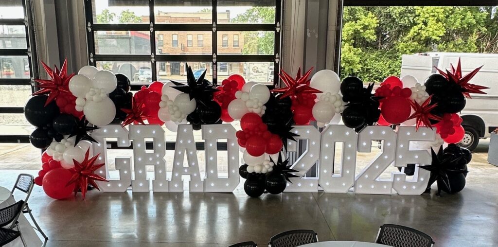 GRAD 2025 light-up marquee letters with red, black, and white organic balloon garland featuring chrome starburst balloons. Indoor graduation party decor by Party Perks in St. Louis. Graduation photo backdrop, custom balloon installation, luxury graduation balloon decor, balloon styling for events, and best graduation balloons in the Greater St. Louis area.