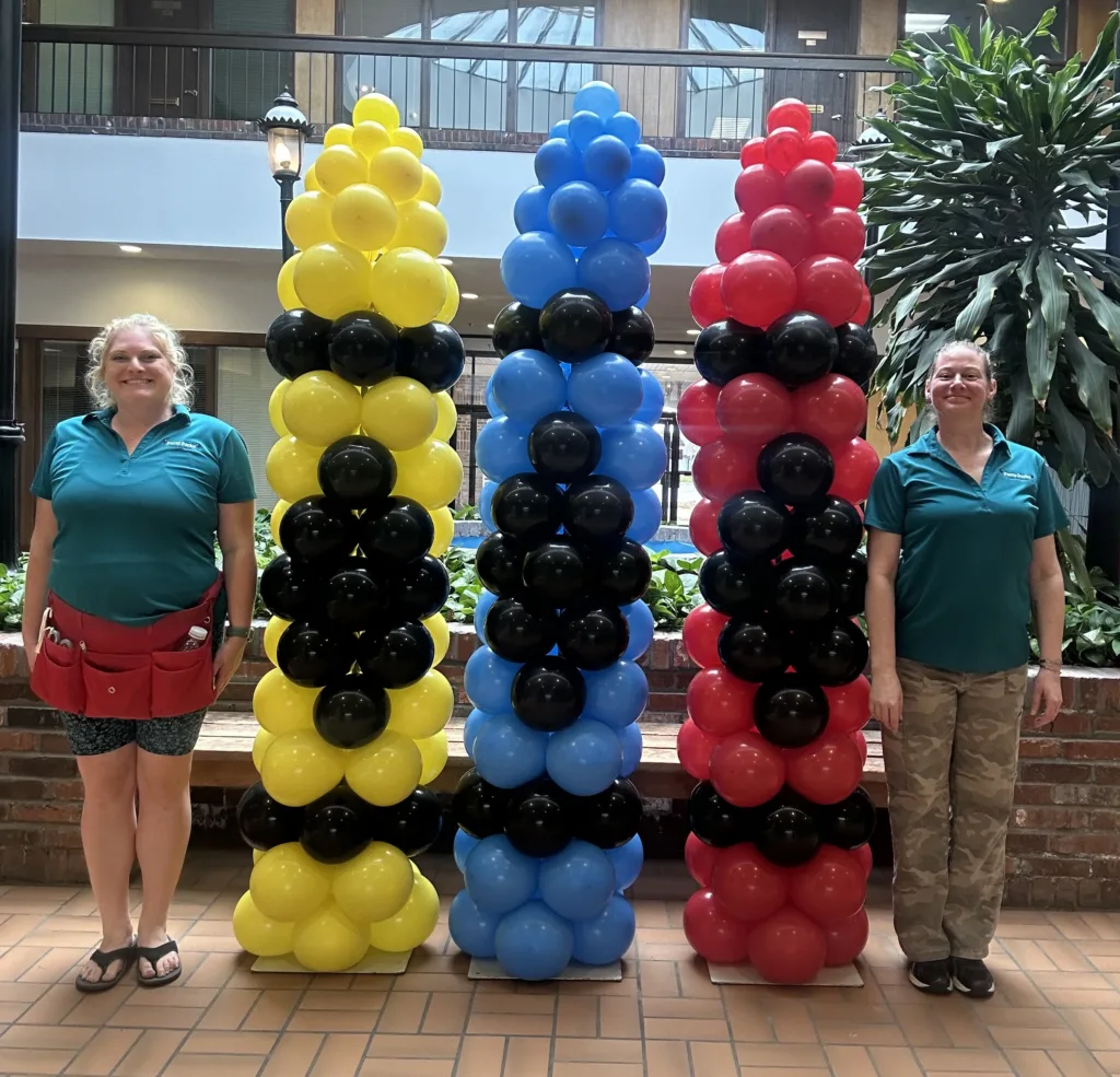 Two balloon artists stand beside three large balloon sculptures designed to look like 7-foot crayons in yellow, blue, and red at a back-to-school event in St. Charles, MO