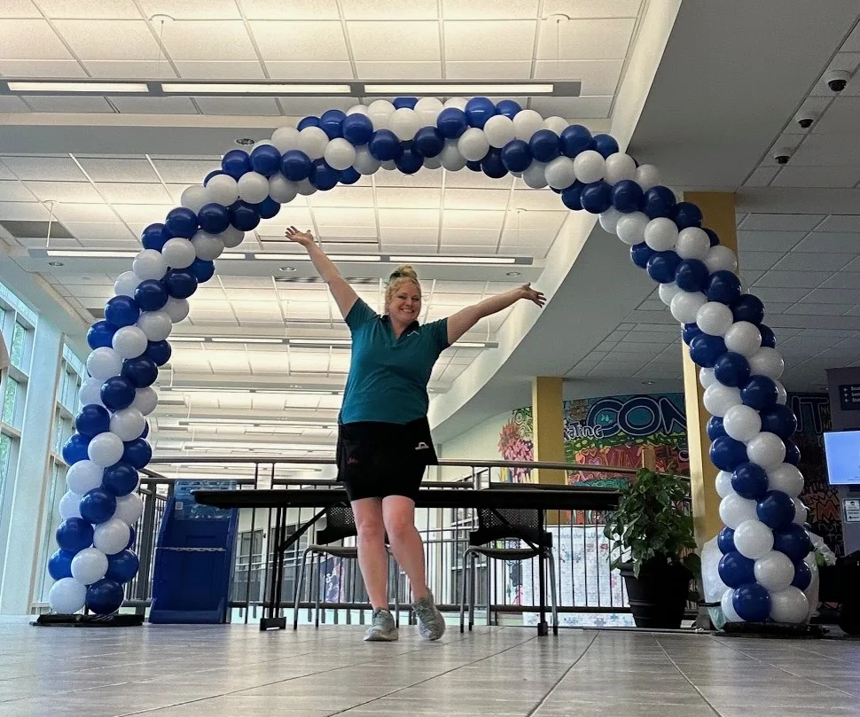 A balloon artist stands with arms raised beneath a classic spiral balloon arch in blue and white at a student orientation at Saint Louis University