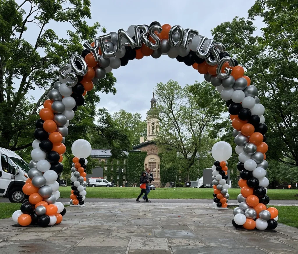 A large outdoor spiral balloon arch with custom silver foil letters reading "50 YEARS OF USG" stands on a college campus, flanked by matching balloon columns in orange, black, white, and silver.