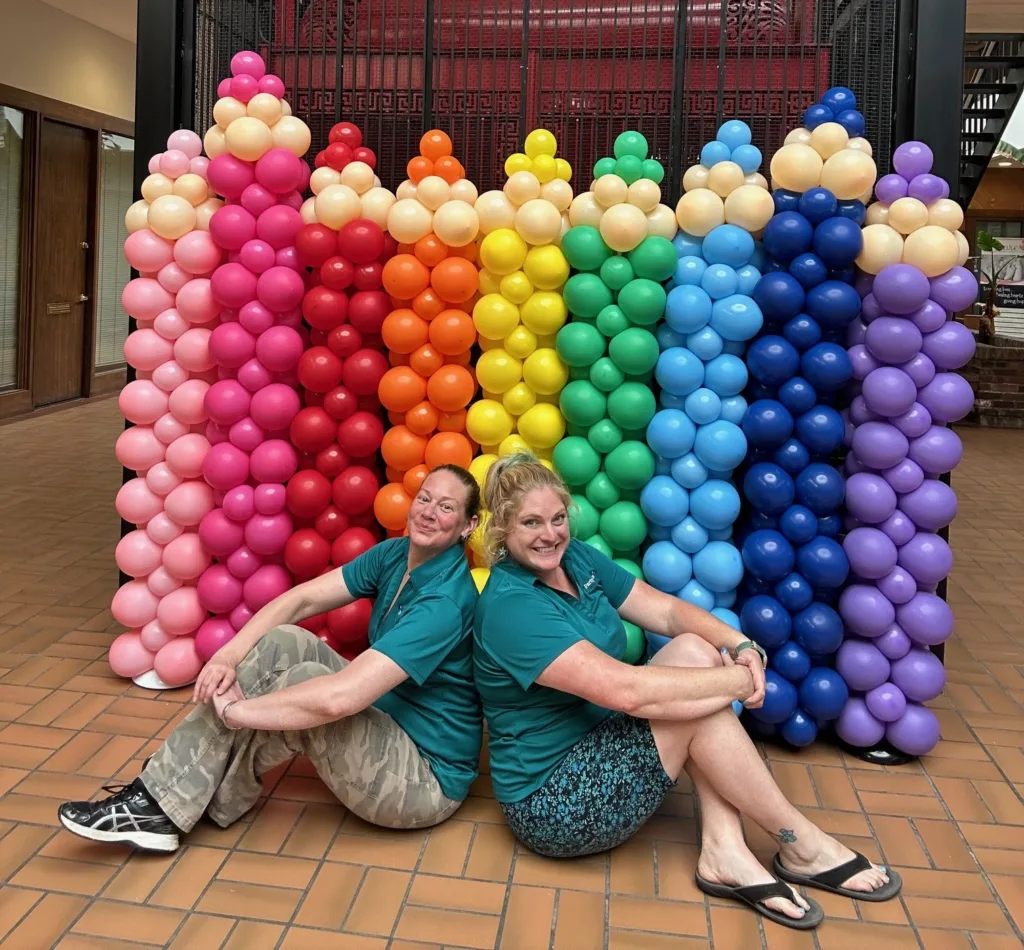 Two women sit smiling in front of a back to school display- rainbow-colored balloon pencil backdrop created from columns of balloons for a back-to-school event in St. Louis.