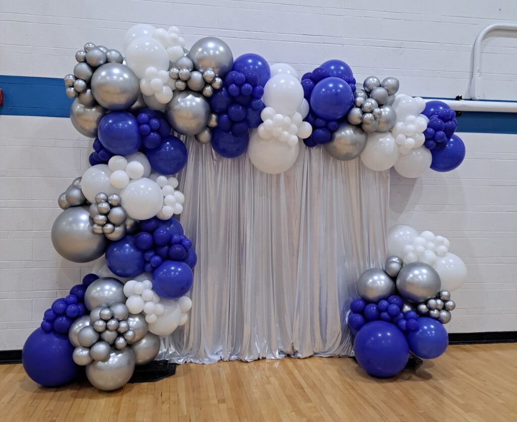 Organic balloon backdrop in blue, white, and silver with layered balloon clusters framing a silver shimmer drape for a photo area at a St. Louis event.