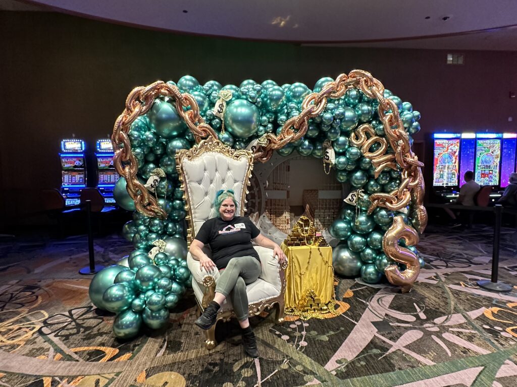 Casino-themed organic balloon installation with metallic teal balloons, giant gold balloon chains, money bag accents, and Jules Perkovic seated in a gold throne chair at a St. Louis casino event.