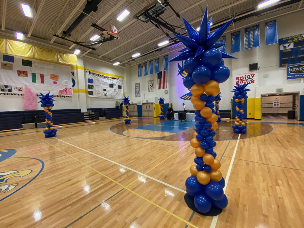 Blue and gold balloon balloon towers with metallic blue starburst toppers arranged around a school gymnasium for a dance or pep rally.