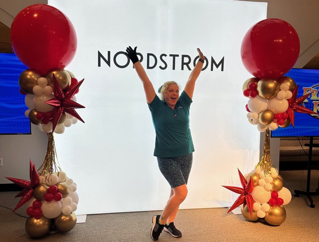 Red, white, and gold balloon columns with metallic starburst accents installed at Nordstrom, with a balloon artist posing in front of a large illuminated Nordstrom backdrop.