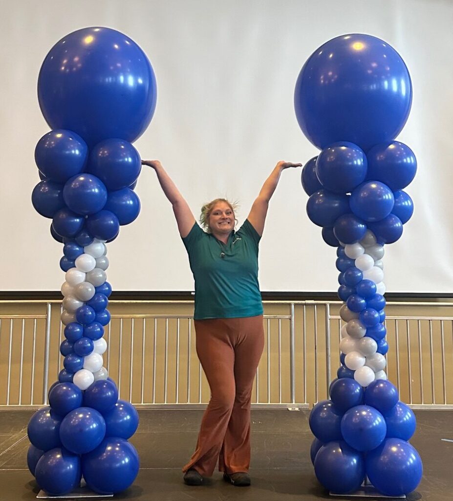Pair of blue and white spiral balloon columns with large blue topper balloons on a stage, with a balloon artist posing between them.