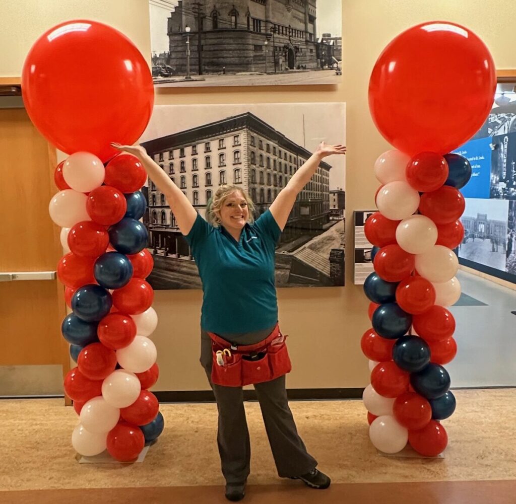 Pair of red, white, and blue spiral balloon columns with large red topper balloons, installed indoors with a balloon artist posing between them.