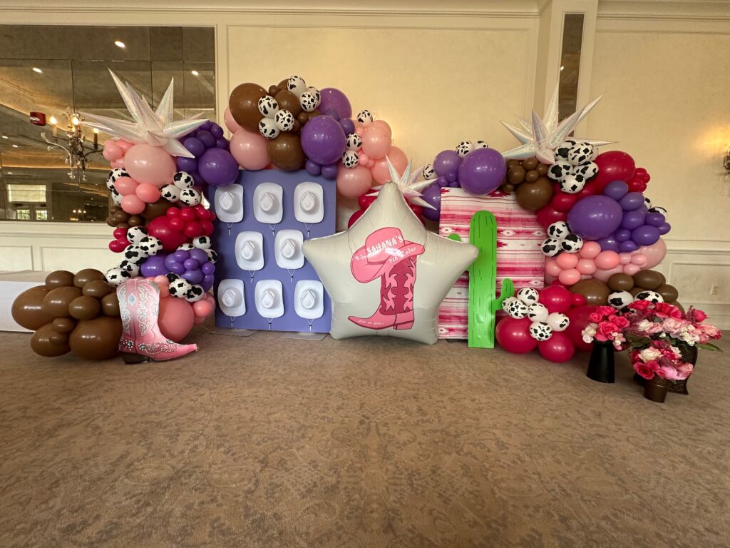 Cowgirl-themed organic balloon backdrop with pink, purple, and brown balloons, cow-print accents, custom vinyl backdrops, cactus cutout, star balloon, and cowgirl boot props for a first birthday celebration.