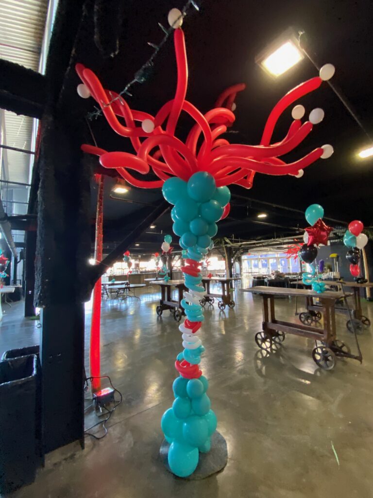 Tall organic balloon tower in teal, red, and white with a dramatic red curly 260s balloon topper, installed inside an industrial-style event venue.