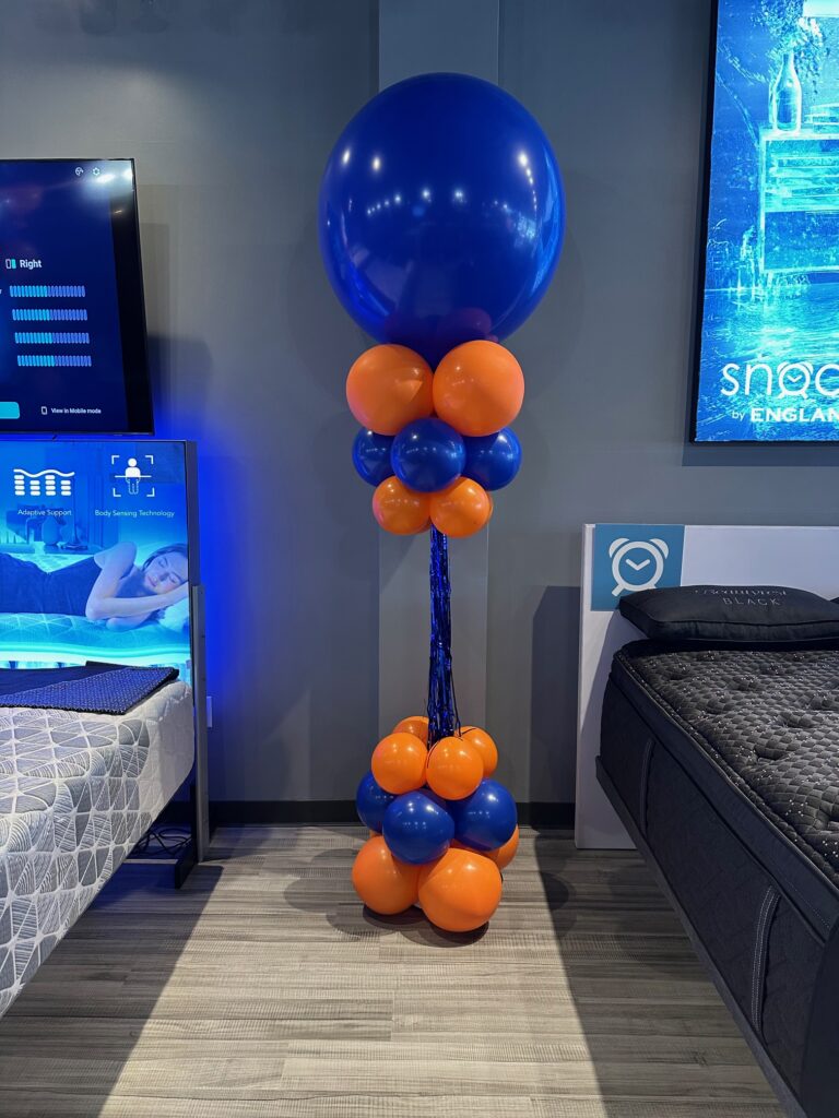 Blue and orange balloon tower with a large blue topper balloon and cascading tinsel center, displayed inside a retail showroom.